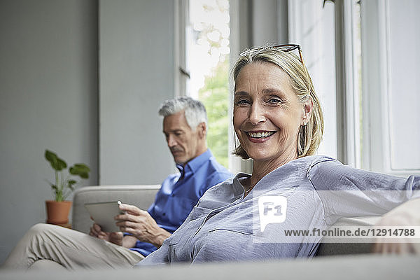Portrait of smiling mature woman on couch with man in background using tablet