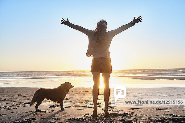 Portugal  Algarve  woman with dog raising arms on the beach at sunset