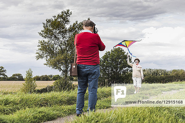 Senior man filming wife flying kite in rural landscape