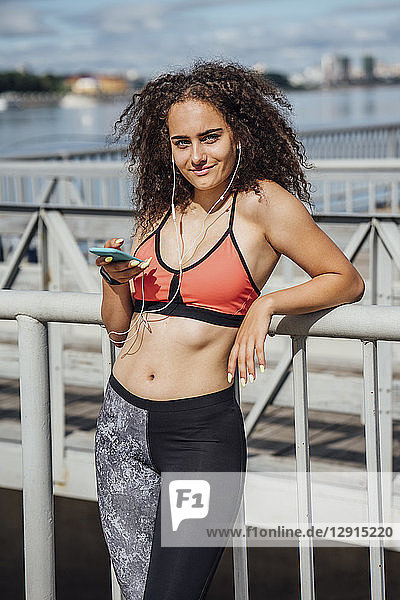 Smiling young athletic woman with smartphone and earbuds at the riverside