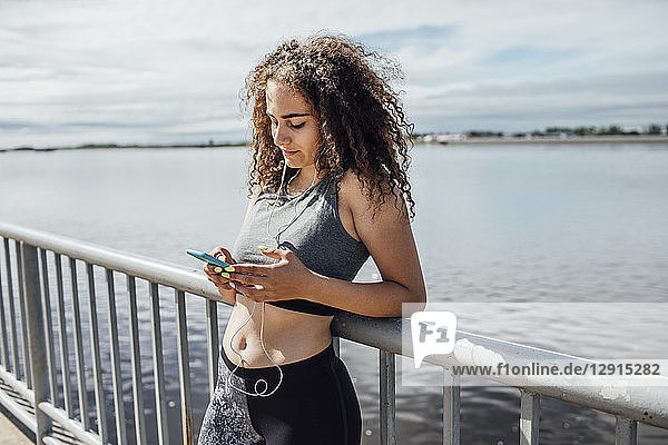 Young athletic woman wearing earbuds and using smartphone at the riverside