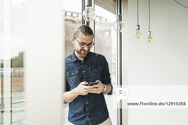 Smiling young businessman using cell phone at the window in office