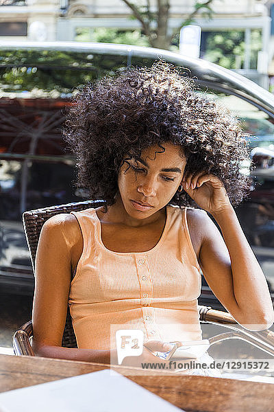 Portrait of young woman with curly hair sitting at sidewalk cafe looking at cell phone