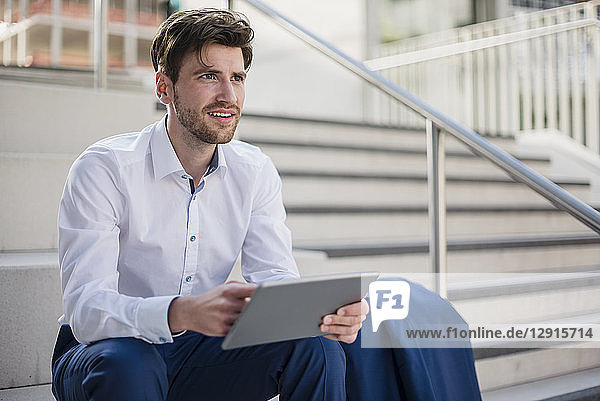 Smiling businessman sitting on stairs in the city with tablet