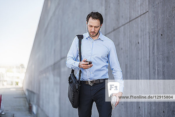 Businessman with earbuds walking along concrete wall looking at smartphone