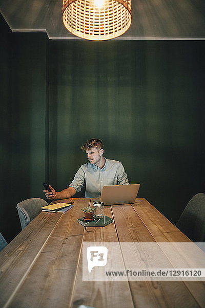 Man sitting in office  working late in his start-up company  using laptop and smartphone