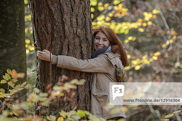 Portrait of happy teenage girl hugging tree trunk in autumnal forest