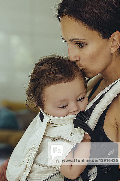 Mother kissing and carrying her baby girl in sling at home