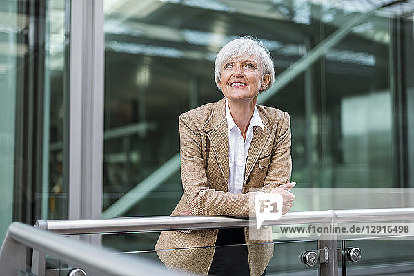 Portrait of smiling senior businesswoman leaning on railing in the city looking up