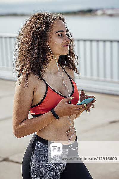 Smiling young athletic woman with smartphone and earbuds at the riverside
