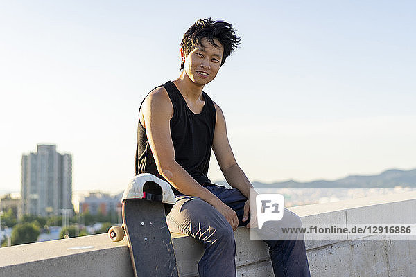 Portrait of smiling young man sitting on wall next to skateboard