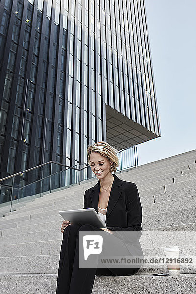 Blond businesswoman with coffee to go sitting on stairs in front of modern office building using tablet