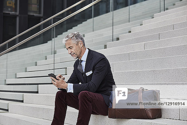 Fashionable businessman with travelling bag sitting on stairs using mobile phone