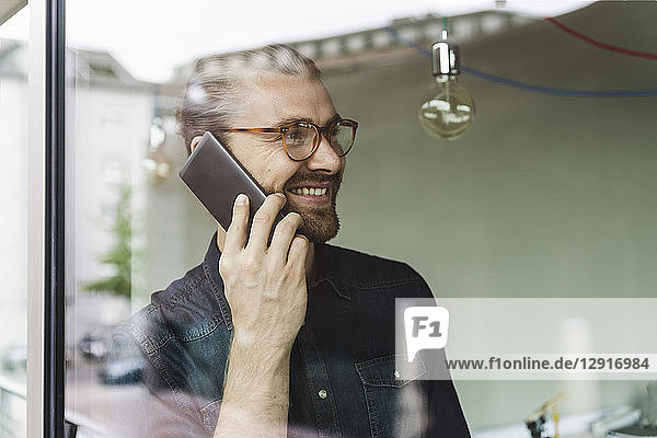 Smiling young businessman on cell phone in office behind windowpane