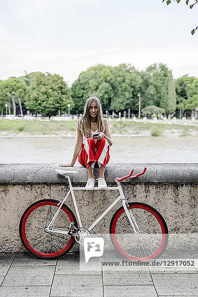 Teenage girl with cell phone sitting at the riverside next to bicycle