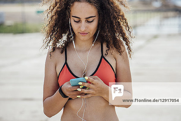 Young athletic woman wearing earbuds and using smartphone