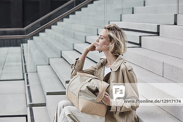 Fashionable blond businesswoman wearing beige trenchcoat sitting on stairs with travelling bag