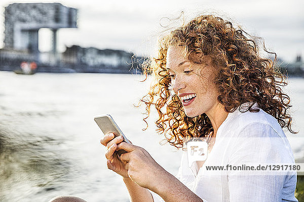 Germany  Cologne  portrait of amazed young woman sitting at riverside looking at cell phone