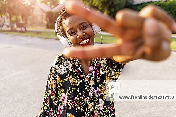 Happy fashionable young woman with headphones outdoors at sunset making victory gesture