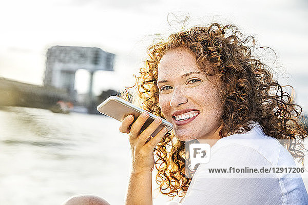 Germany  Cologne  portrait of happy young woman on the phone sitting at riverside in the evening