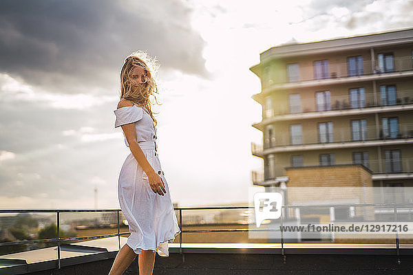 Blond young woman standing on roof terrace at sunset wearing white dress