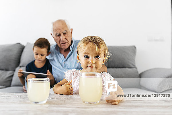 Portrait of little girl with glass of lemonade at home with brother and grandfather in the background
