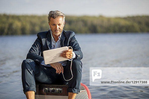 Portrait of relaxed businessman with earphones and tablet sitting on paddleboard at lake