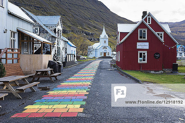 Rainbow colored brick road between houses leading to small church at edge of town Seydisfjordur Iceland