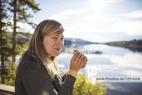 Side view of smiling woman looking at camera while eating hot dog on lakeshore