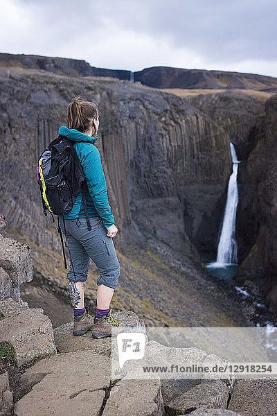 Weibliche Rucksacktouristin steht auf einem Felsen und blickt auf den Litlanesfoss-Wasserfall in der Ferne  Island
