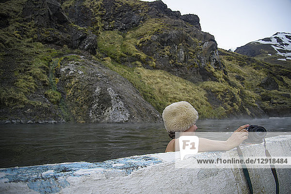 Frau mit Pelzmütze und Kamera in heißer Quelle in natürlicher Umgebung  Seljavallalaug  Island