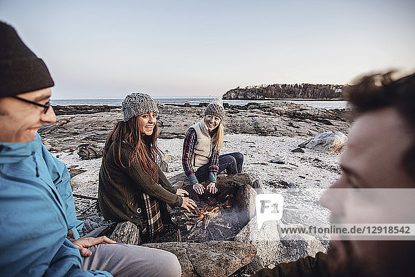 Vier erwachsene Freunde  die sich am Lagerfeuer unterhalten und lächeln  während sie an einem felsigen Küstenstrand im Herbst campen  Portland  Maine  USA