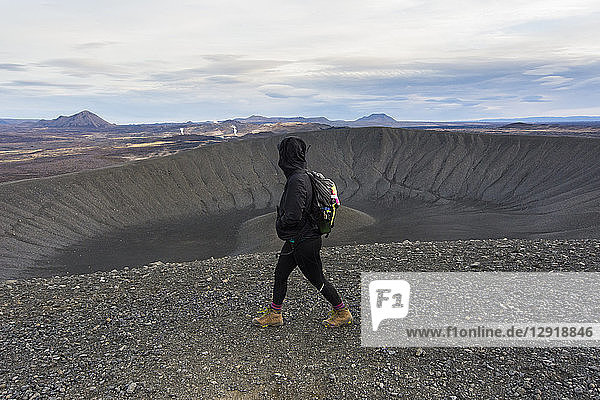 Eine junge  schwarz gekleidete Frau in Stiefeln geht auf dem Kamm des Hverfjall-Kraters entlang  während im vulkanischen Hintergrund Dampf aufsteigt.