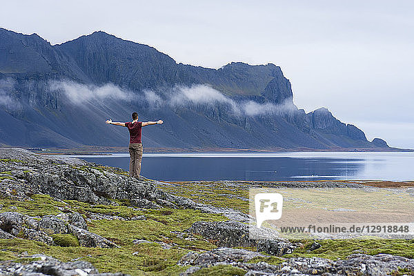 A young man wearing a purple t-shirt stands on top of a moss covered rock as he opens his arms and admires the mountainous coastline of East Iceland Hringvegur East Iceland Iceland