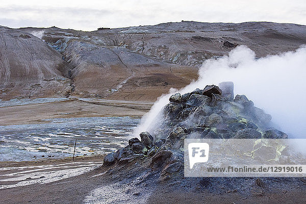 Eine aktive Dampfquelle spuckt Schwefel und andere giftige Gase in Form von Dämpfen aus  die den Boden in der Umgebung blau und leuchtend gelb gefärbt haben. Im Hintergrund ist der Berg Namafjall im Hverarˆšˆ'ndor Hverir Geothermalgebiet am Namaskard-Pass in Nordisland zu sehen.