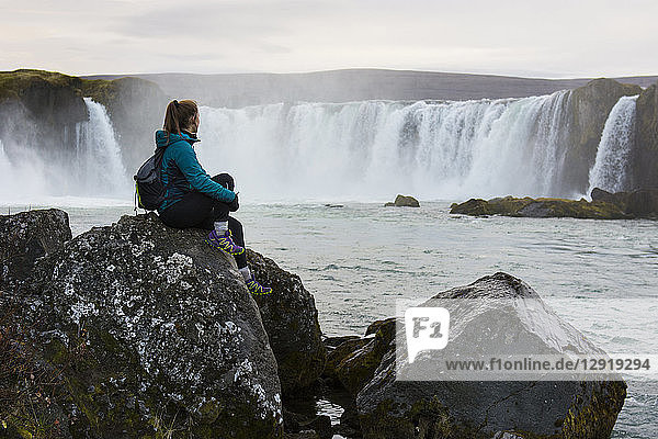 Porträt einer Wanderin  die auf einem Felsen sitzt und den plätschernden Godafoss-Wasserfall betrachtet  Island