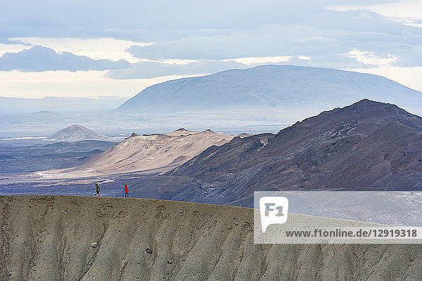 Zwei Personen in farbenfroher Kleidung spazieren entlang des Kammes des Viti-Kraters  mit einer bergigen Landschaft im Hintergrund  im Vulkangebiet Krafla  in der Nähe des Myvatn-Sees in Nordisland.
