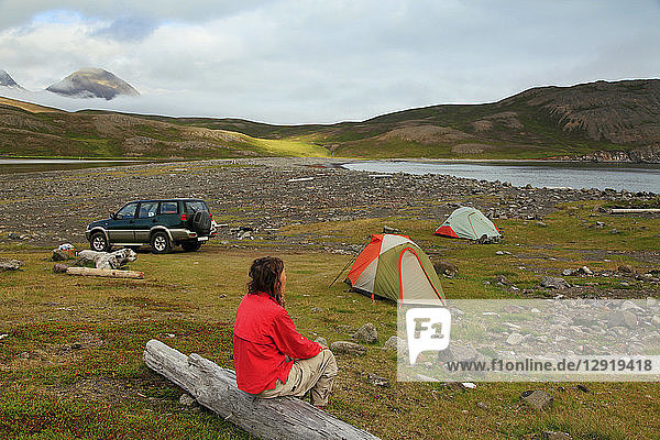 Side view shot of single mature woman sitting on log near camping tents and 4x4 car  Iceland