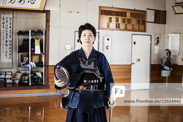 Japanische Kendo-Kämpferin steht in einer Turnhalle  hält Kendo-Maske und Schwert und schaut in die Kamera.