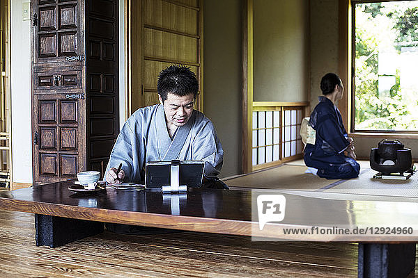Japanischer Mann in Kimono  der in einem traditionellen japanischen Haus auf dem Boden sitzt und auf ein digitales Tablett schaut.