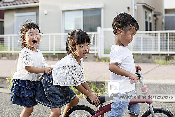Porträt von zwei japanischen Mädchen und einem Jungen  die mit einem Fahrrad auf der Straße spielen und in die Kamera lächeln.