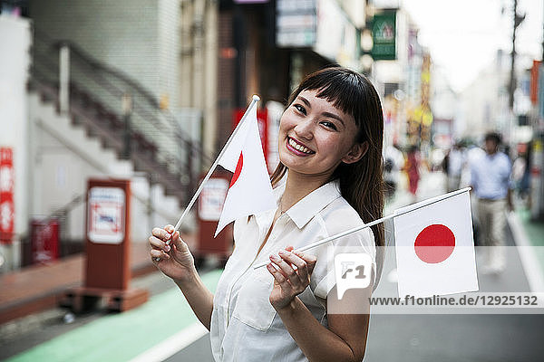 Smiling Japanese woman with long brown hair wearing white short-sleeved blouse standing in a street  holding small Japanese flag.