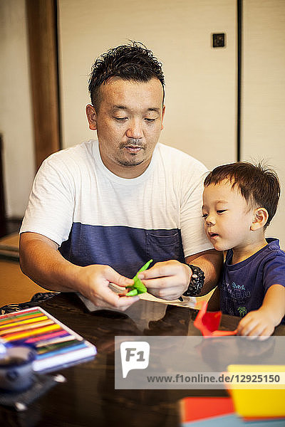 Japanischer Mann und kleiner Junge sitzen an einem Tisch und basteln Origami-Tiere aus buntem Papier.