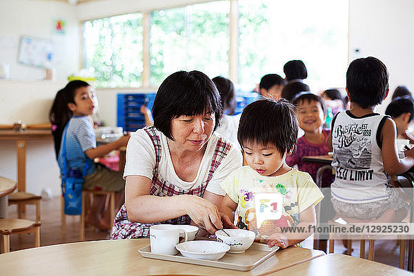 Eine Lehrerin sitzt in einer japanischen Vorschule am Tisch und hilft dem Jungen beim Mittagessen.