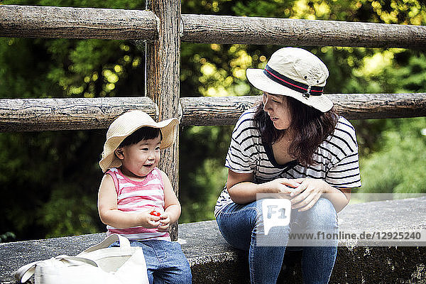 Lächelnde Japanerin sitzt neben einem kleinen Mädchen mit Sonnenhut  gestreiftem Oberteil und Jeans.