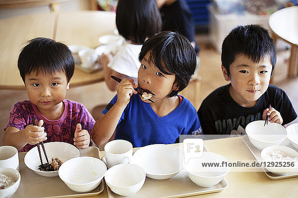 High angle view of three boys sitting at a table in a Japanese preschool eating with chopsticks.