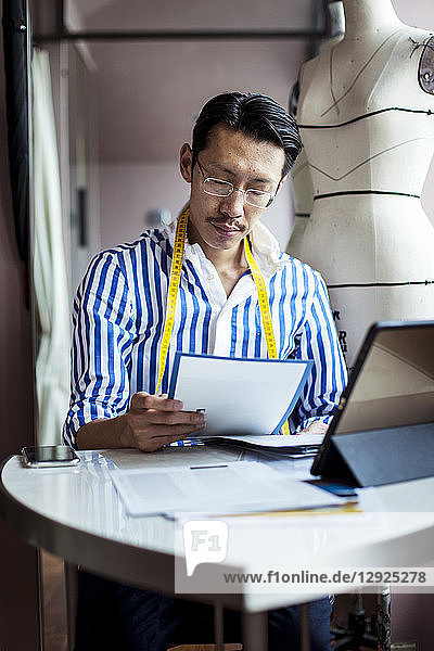 Japanischer männlicher Modedesigner  der in seinem Atelier arbeitet  am Tisch sitzt und auf ein digitales Tablett schaut.