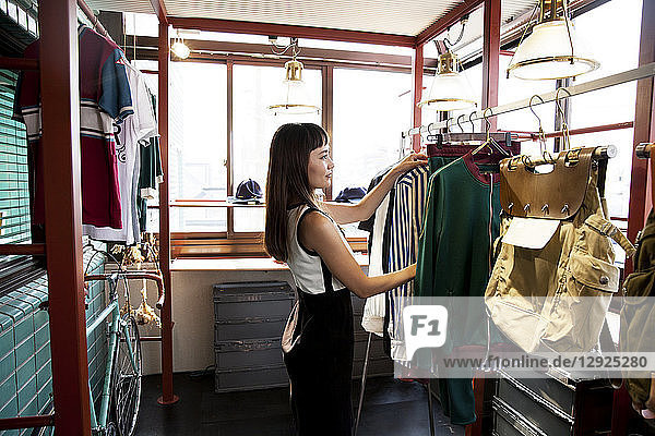 Japanische Verkäuferin  die in einem Bekleidungsgeschäft steht und Kleider auf einer Schiene arrangiert.