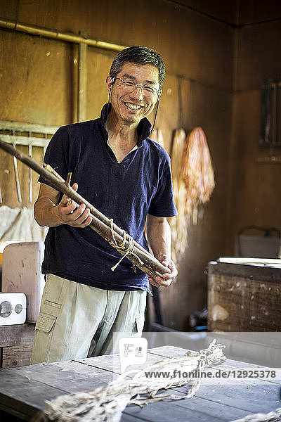 Japanese man standing in a traditional Washi workshop  holding a bunch of mulberry twigs  plant fibre ingredients and strips of fibre on a table.