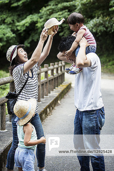 Japanese girl  smiling woman holding hat and man carrying toddler on his shoulders standing on wooden bridge.